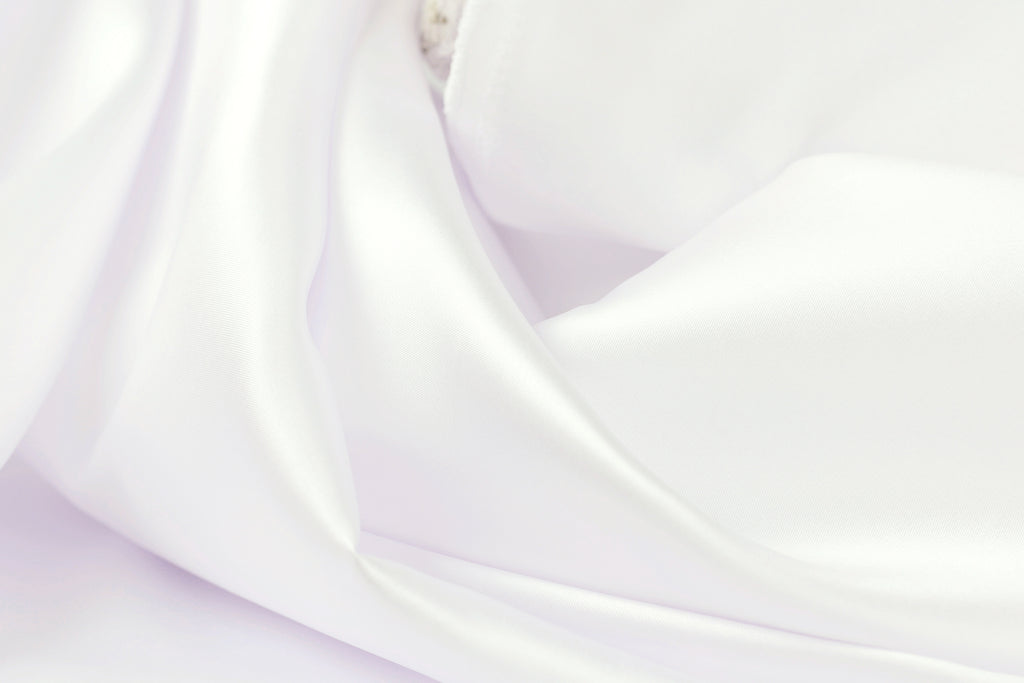 The image is a close-up of a soft, white fabric, possibly silk or satin, with a smooth and flowing texture.