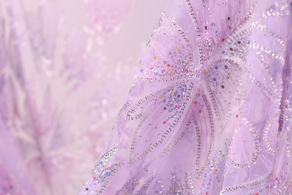 The image is a close-up of a delicate, intricately embroidered fabric with a soft, pastel pink hue. The fabric features a floral pattern with small, colorful beads or sequins scattered throughout, adding a touch of sparkle and detail to the overall design.