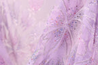 The image is a close-up of a delicate, intricately embroidered fabric with a soft, pastel pink hue. The fabric features a floral pattern with small, colorful beads or sequins scattered throughout, adding a touch of sparkle and detail to the overall design.