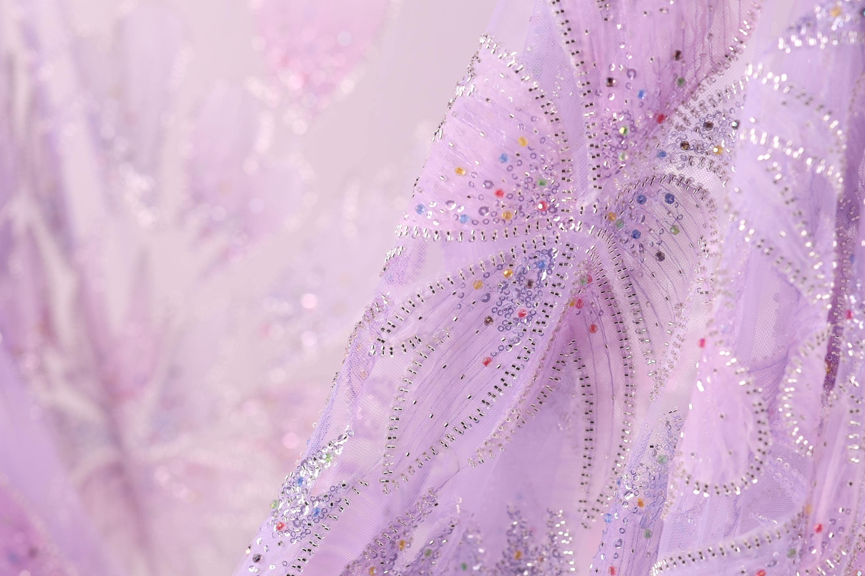 The image is a close-up of a delicate, intricately embroidered fabric with a soft, pastel pink hue. The fabric features a floral pattern with small, colorful beads or sequins scattered throughout, adding a touch of sparkle and detail to the overall design.