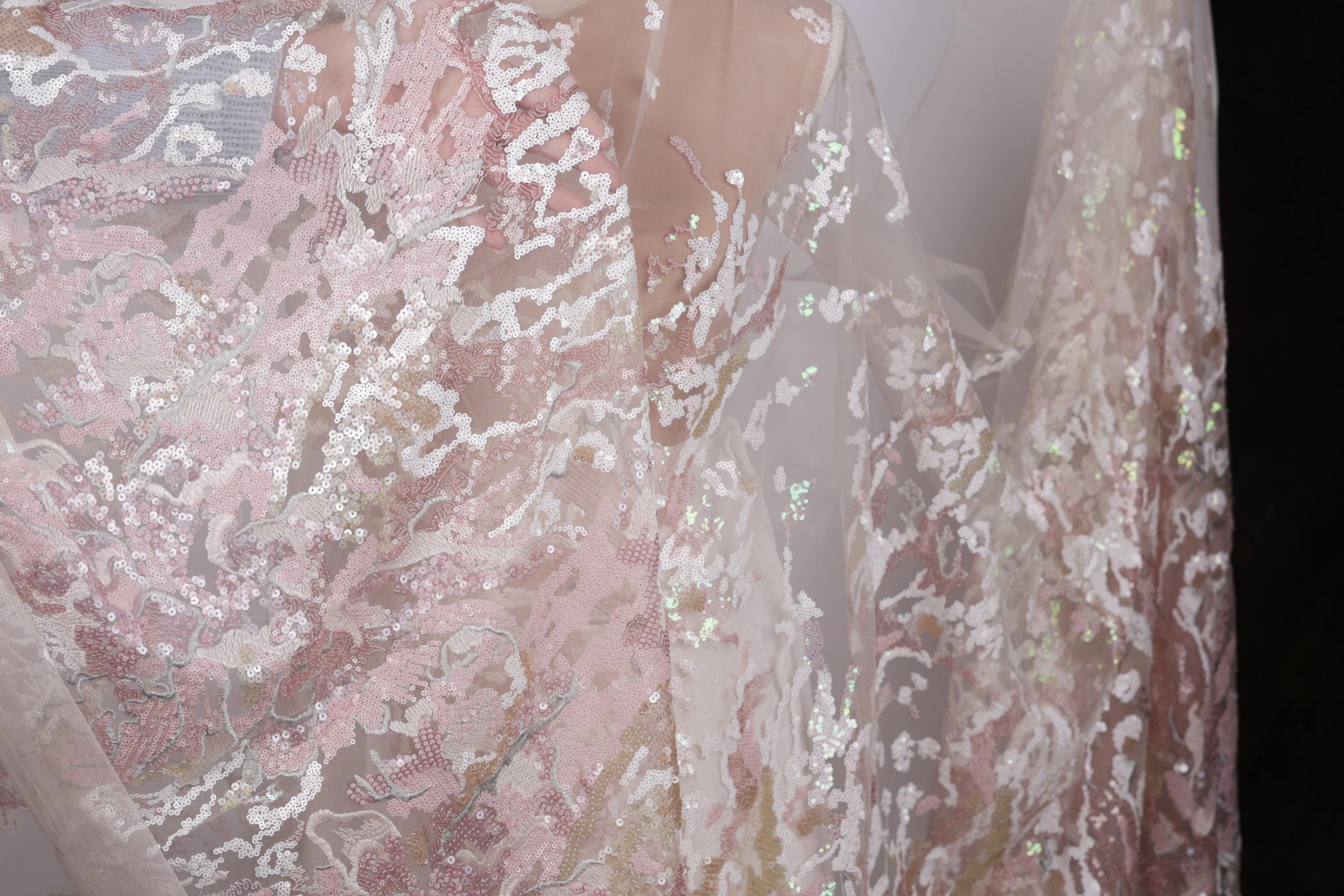 a close-up view of a delicate, lace-like fabric with a floral pattern in shades of pink and white. The fabric appears to be made of sheer material, giving it a delicate and airy appearance.