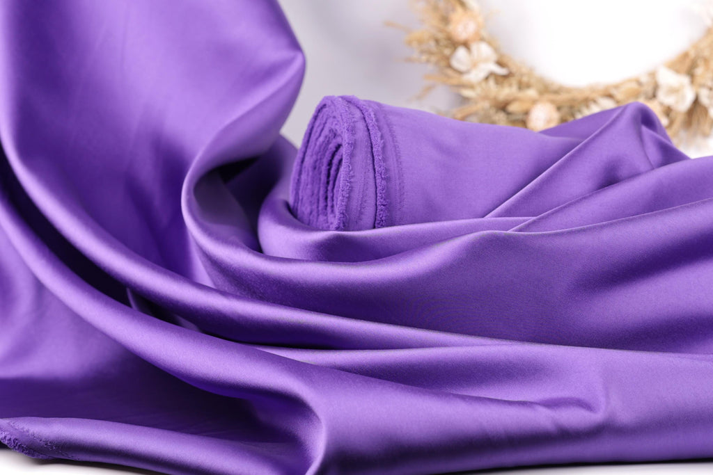 A close-up view of a purple satin fabric, with a wreath of dried flowers visible in the background.