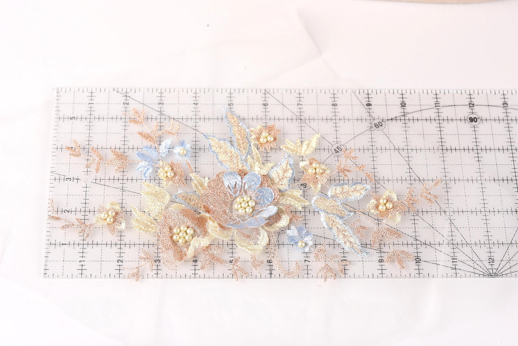 a close-up view of a delicate floral embroidery on a transparent ruler. The embroidery is composed of various shades of blue and peach flowers, with small white pearls scattered throughout the design. The ruler serves as a guide for the embroidery artist, helping them maintain the precise measurements and proportions of the floral pattern.