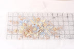 a close-up view of a delicate floral embroidery on a transparent ruler. The embroidery is composed of various shades of blue and peach flowers, with small white pearls scattered throughout the design. The ruler serves as a guide for the embroidery artist, helping them maintain the precise measurements and proportions of the floral pattern.