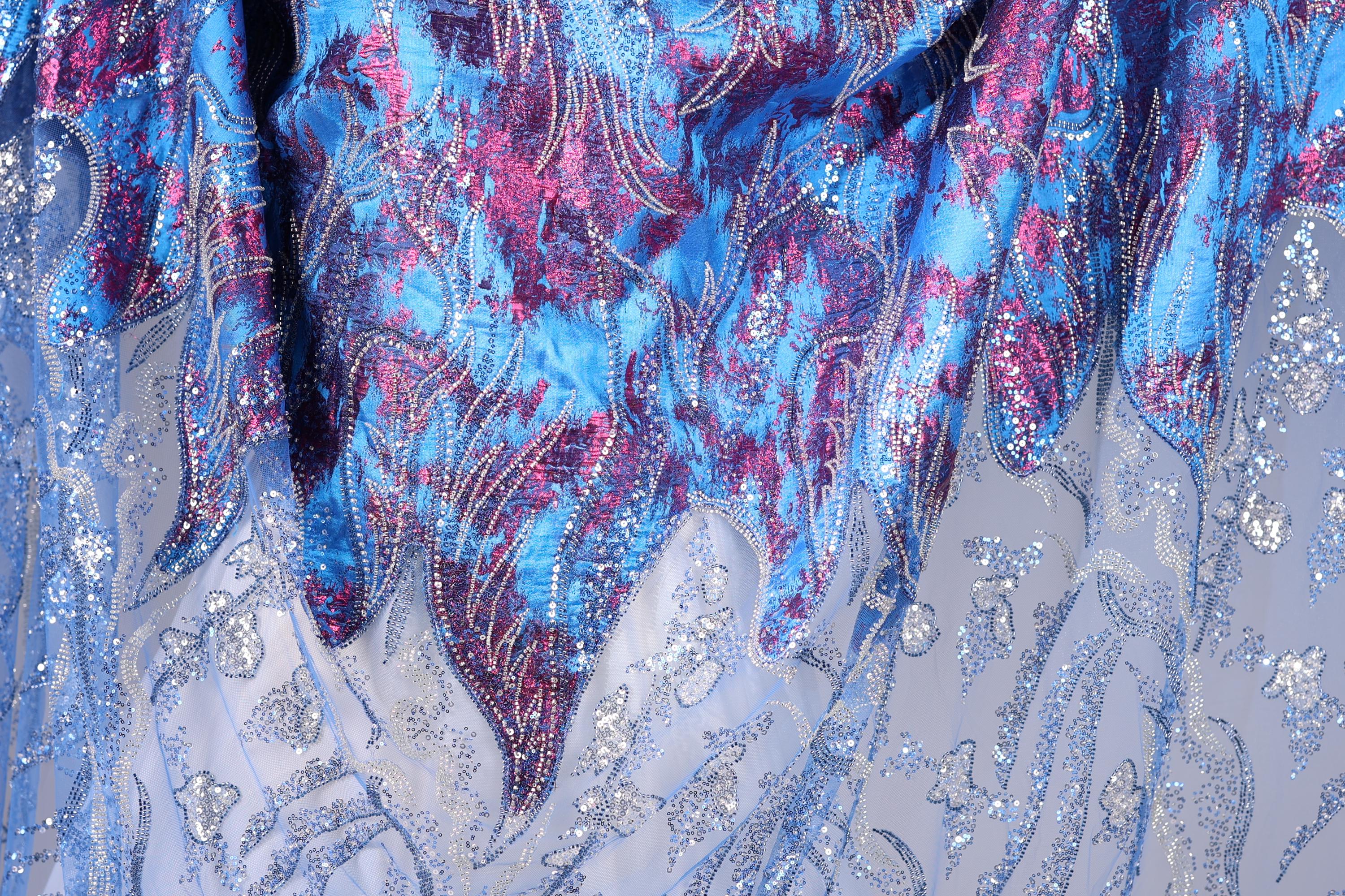 a close-up view of a fabric with a vibrant, multicolored pattern. The fabric appears to be a blend of blue, purple, and pink hues, with intricate designs and textures that create a visually striking and detailed appearance.