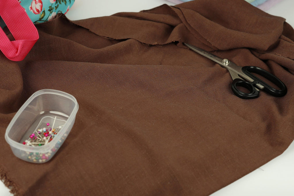 a brown fabric, a pair of black scissors, a clear plastic container with pins, and a red object in the background.