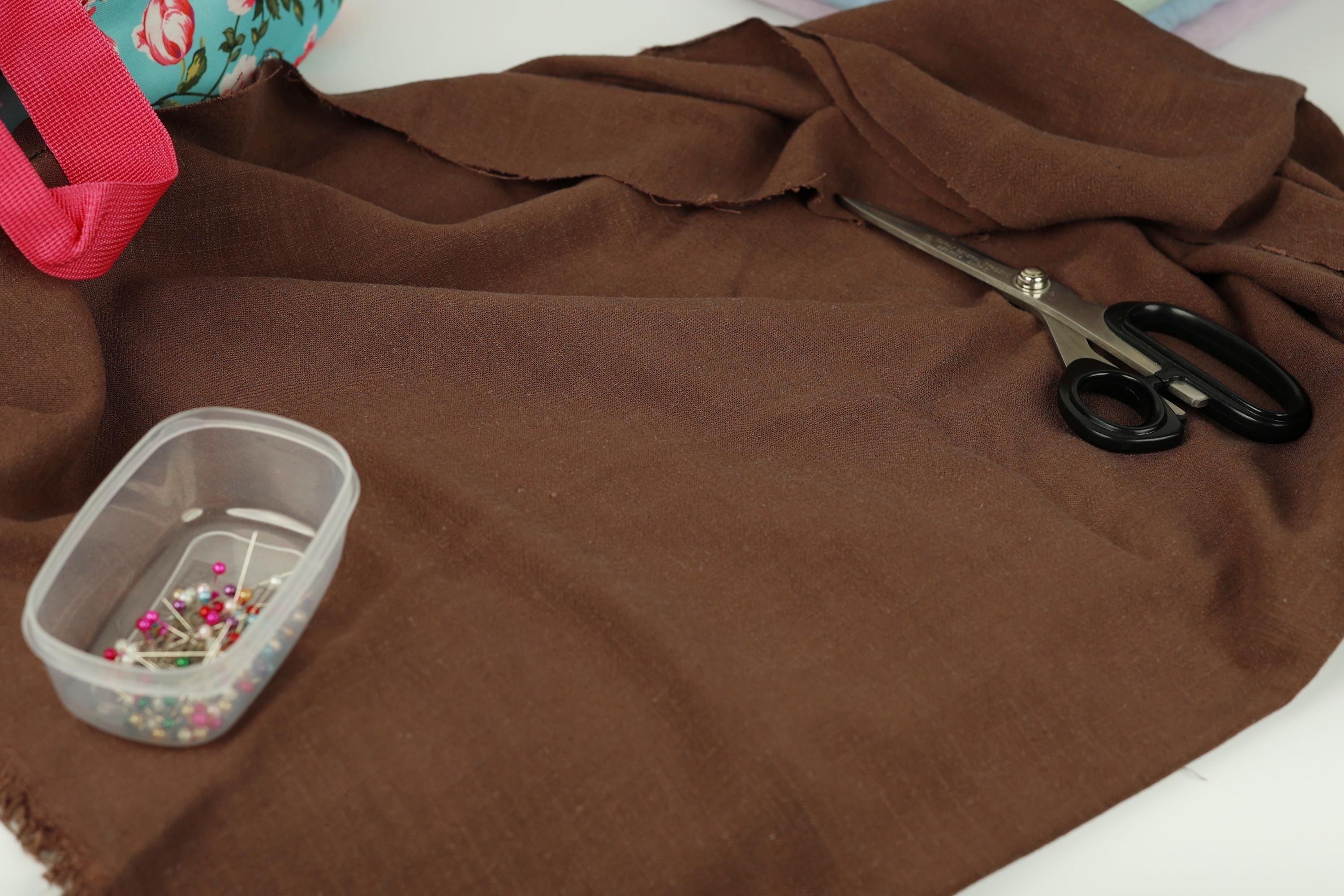 a brown fabric, a pair of black scissors, a clear plastic container with pins, and a red object in the background.