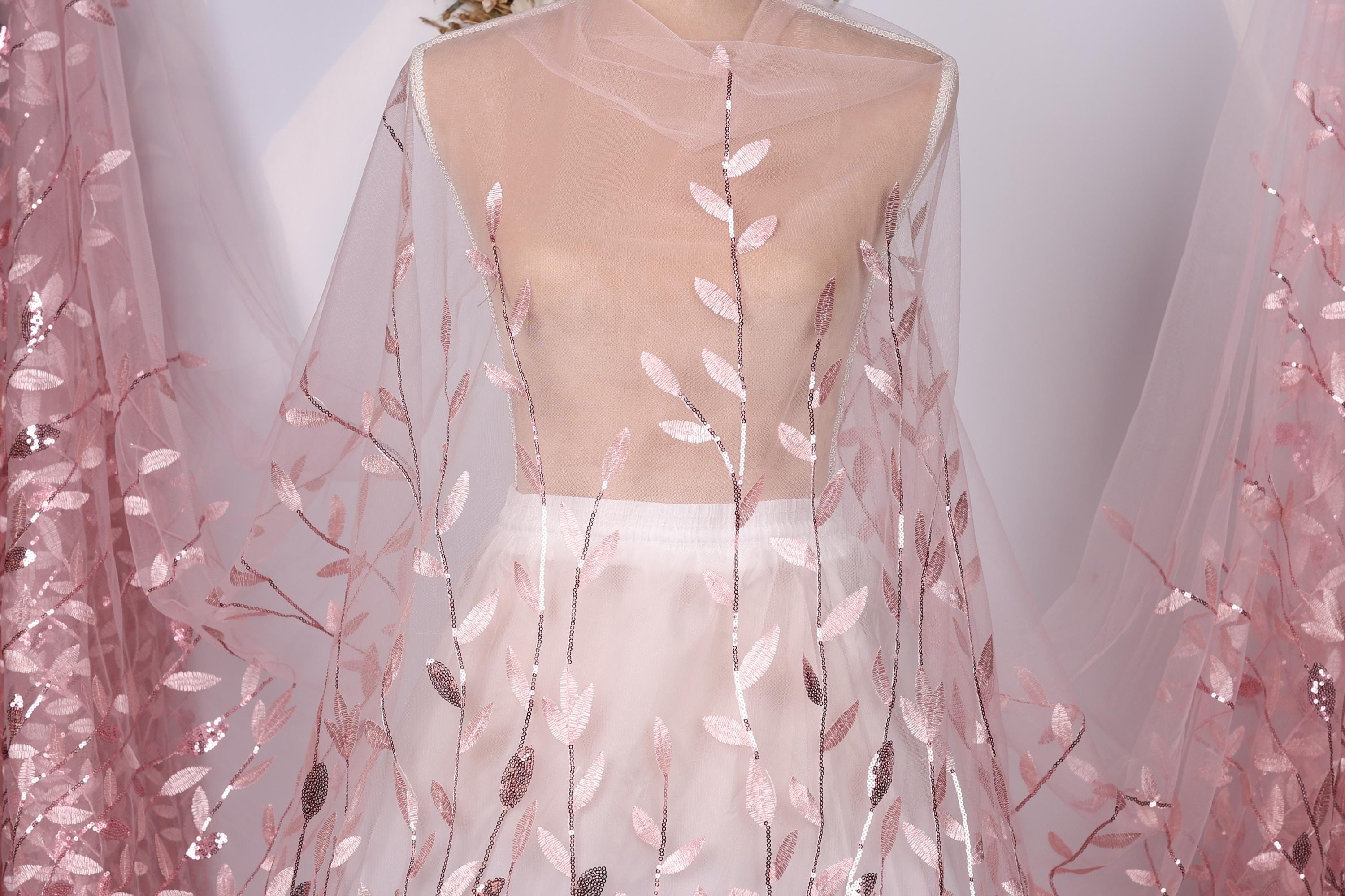 A mannequin wearing a white dress with pink floral embroidery on the skirt and a sheer, transparent fabric draped over the body.