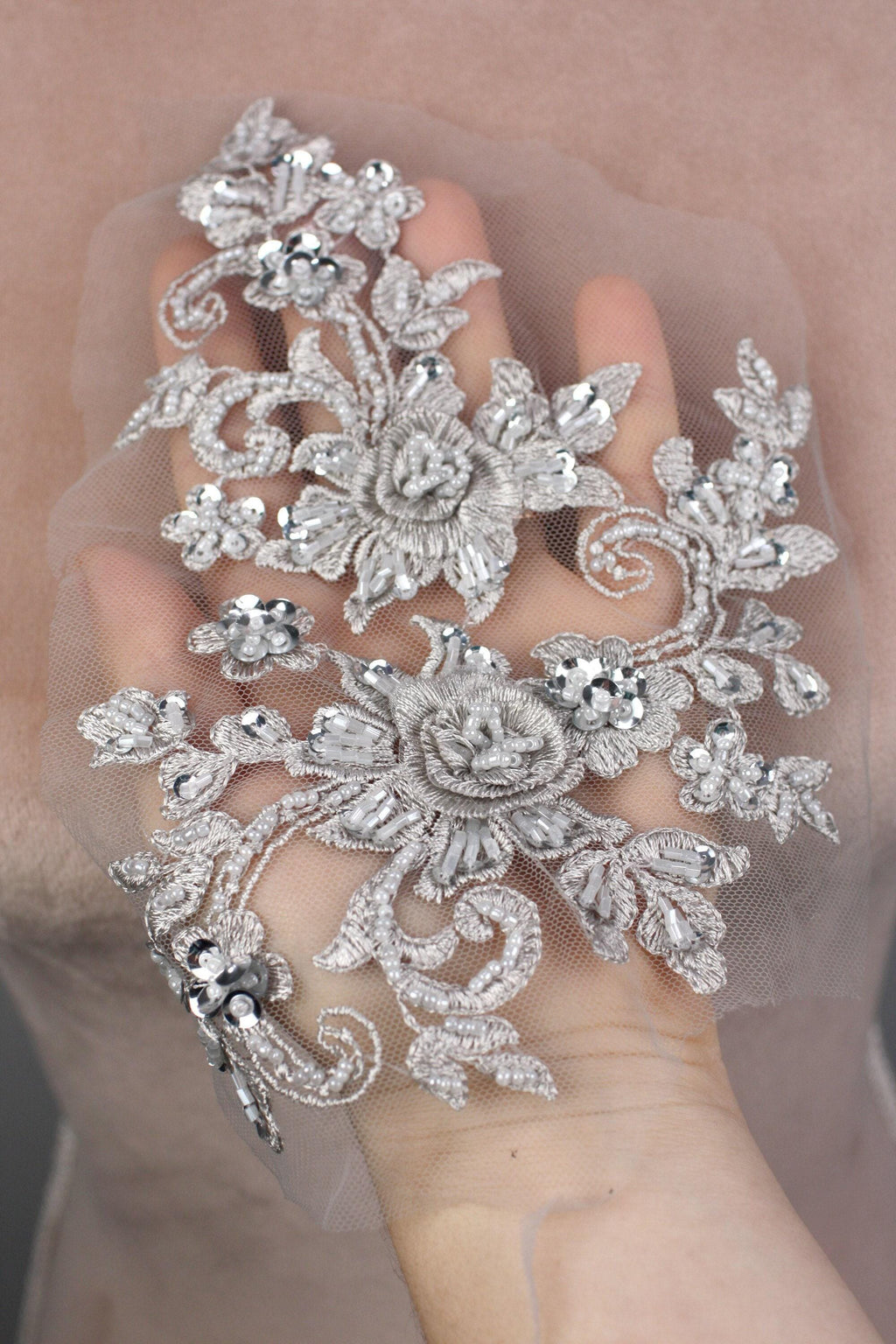 A hand holding a delicate, ornate piece of lace or fabric adorned with intricate floral and leaf designs, embellished with sparkling rhinestones or crystals.