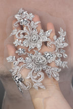 A hand holding a delicate, ornate piece of lace or fabric adorned with intricate floral and leaf designs, embellished with sparkling rhinestones or crystals.