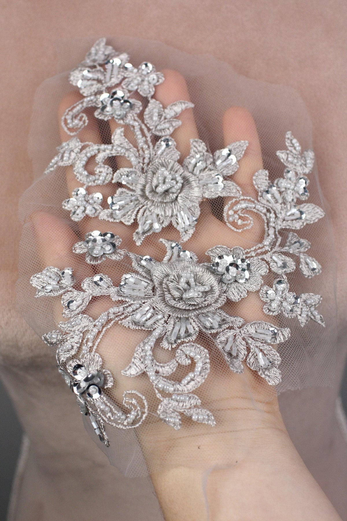 A hand holding a delicate, ornate piece of lace or fabric adorned with intricate floral and leaf designs, embellished with sparkling rhinestones or crystals.