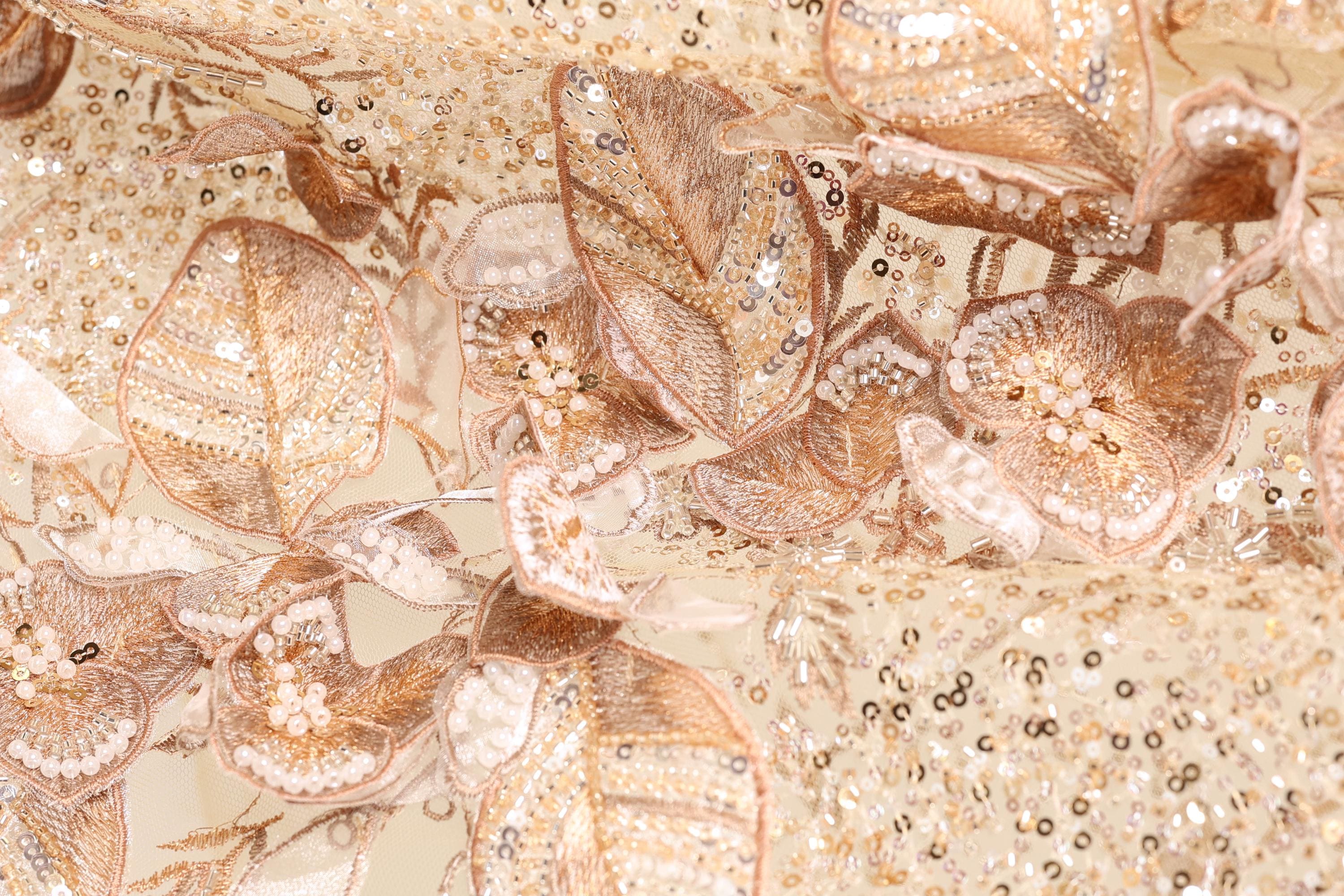 The image is a close-up of a fabric with intricate gold and white embroidered designs, including flowers and leaves. The fabric appears to be a delicate and ornate material, possibly used for clothing or other decorative purposes.