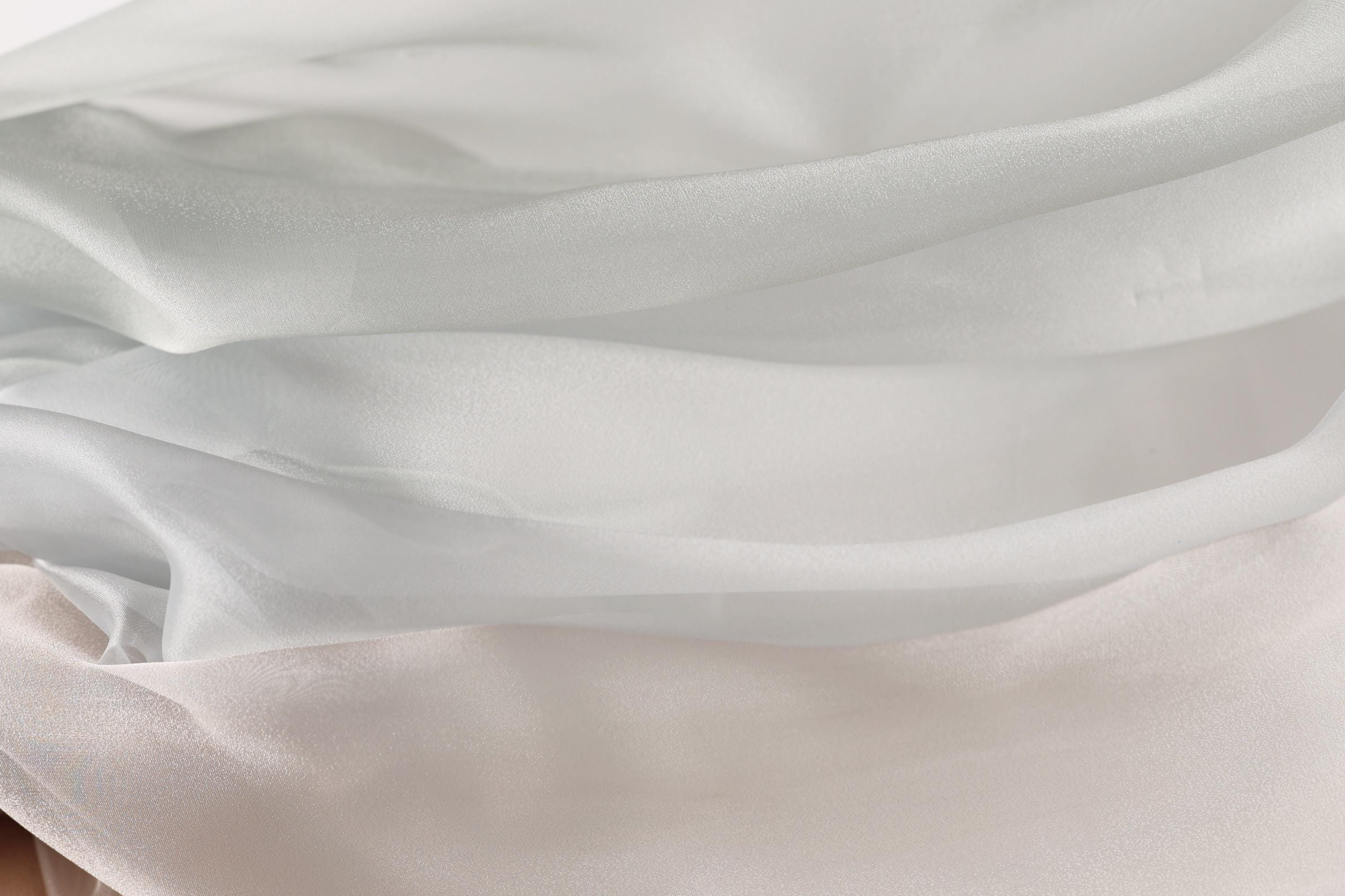The image is a close-up of a soft, flowing fabric with a gradient of white and light gray colors. The fabric appears to be made of silk or a similar material, and it has a smooth, shiny texture. The colors are subtle, with the white and light gray shades blending together to create a soft, delicate appearance.