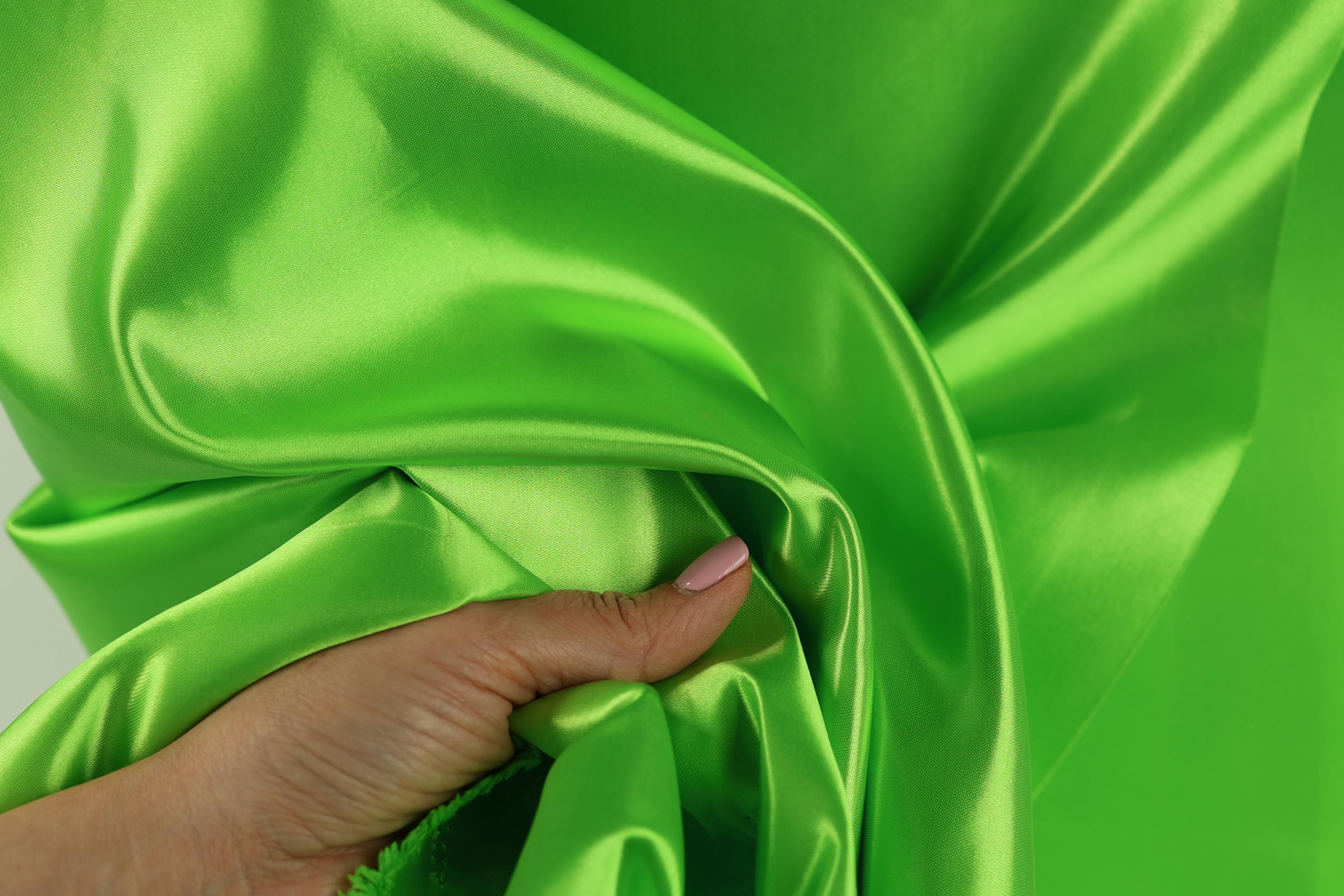 A hand is holding a piece of bright green satin fabric, with the fabric's smooth and shiny texture visible.