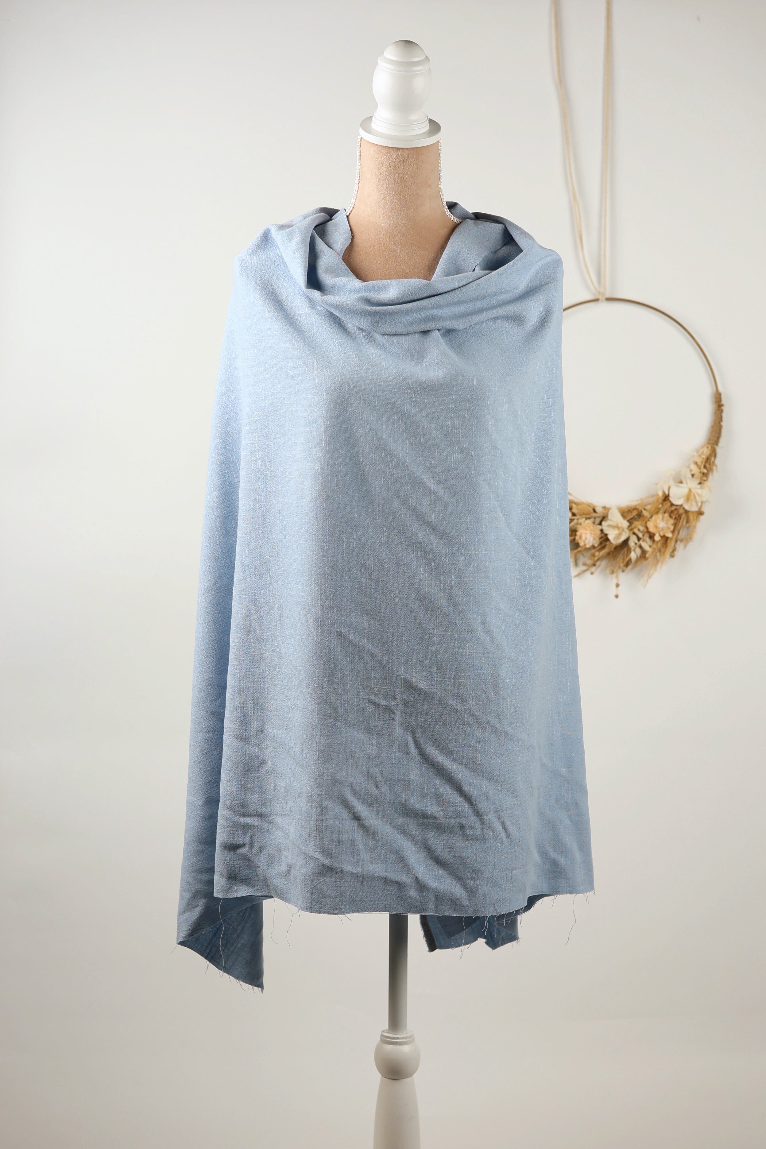 A light blue draped garment is displayed on a mannequin, with a wreath hanging on the wall behind it.