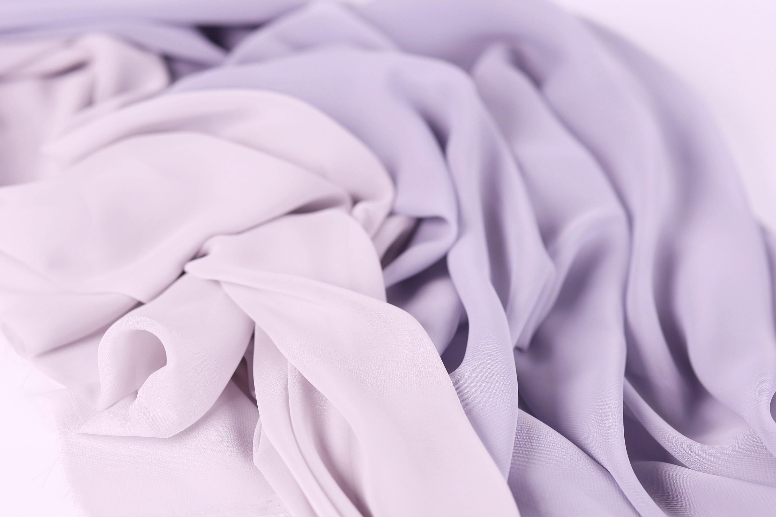 The image is a close-up of a soft, flowing fabric in shades of purple and pink, with a slightly crumpled texture.