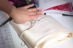 A hand is holding a silver necklace with a pendant, resting on a white fabric, with a pair of scissors and a patterned fabric visible in the background.