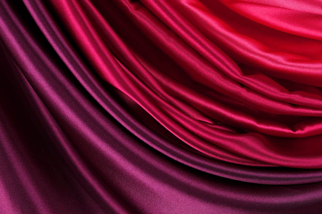 a close-up view of a fabric with a gradient of colors, transitioning from a deep red to a lighter purple. The fabric appears to be made of satin or a similar smooth, shiny material, and it has a flowing, draped appearance.