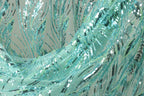 The image is a close-up of a fabric with a shimmering, sequined pattern in shades of blue and green. The fabric appears to be made of a sheer, sheer-like material, giving it a delicate and ethereal appearance. The intricate design and the shimmering sequins create a visually striking and captivating image.
