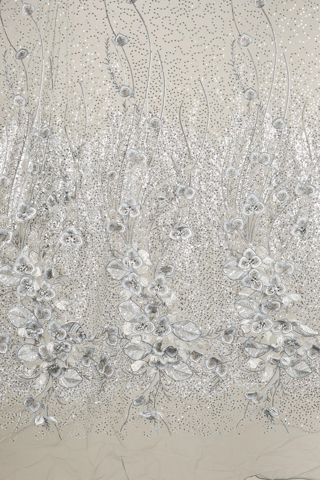a close-up view of a fabric with a floral pattern. The fabric appears to be made of a shiny, metallic material, giving it a metallic, glittery appearance. The pattern consists of numerous small, white flowers or petals arranged in a repeating, intricate design. The fabric is displayed against a light background, which further accentuates the metallic sheen and the intricate details of the floral pattern.