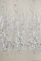 a close-up view of a fabric with a floral pattern. The fabric appears to be made of a shiny, metallic material, giving it a metallic, glittery appearance. The pattern consists of numerous small, white flowers or petals arranged in a repeating, intricate design. The fabric is displayed against a light background, which further accentuates the metallic sheen and the intricate details of the floral pattern.