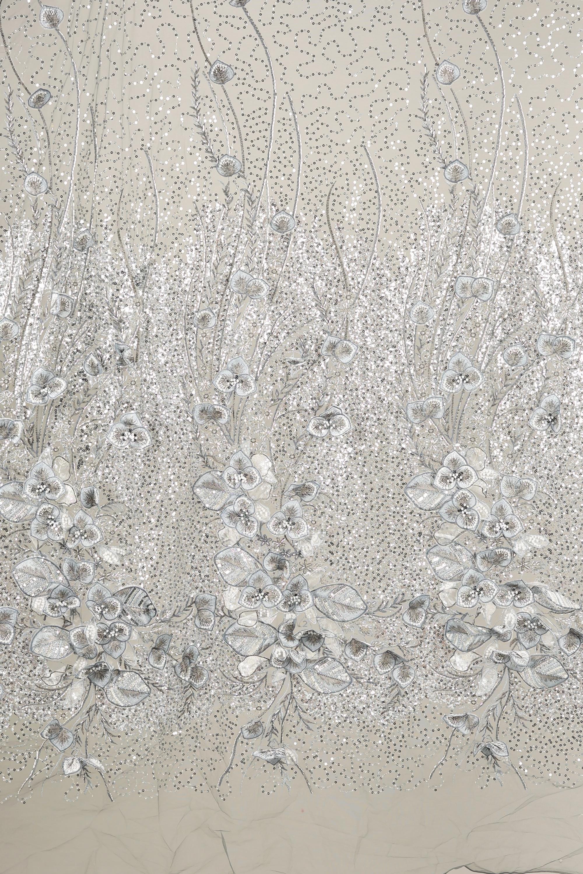 a close-up view of a fabric with a floral pattern. The fabric appears to be made of a shiny, metallic material, giving it a metallic, glittery appearance. The pattern consists of numerous small, white flowers or petals arranged in a repeating, intricate design. The fabric is displayed against a light background, which further accentuates the metallic sheen and the intricate details of the floral pattern.