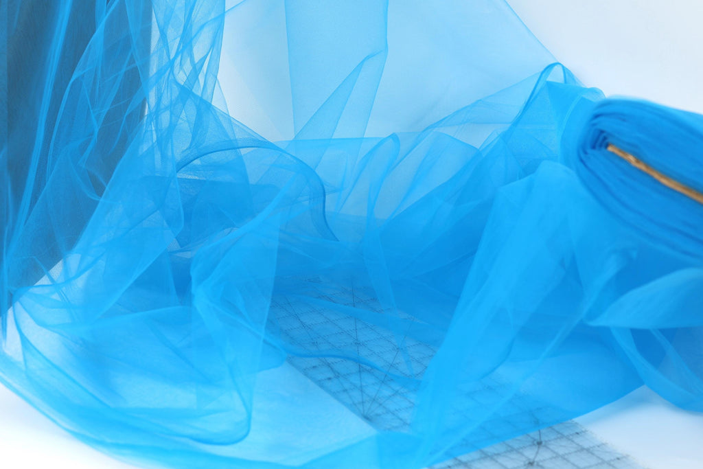 a close-up view of a blue, sheer fabric or chiffon material, with a grid pattern visible underneath. The fabric appears to be folded or crumpled, creating a textured and flowing appearance.