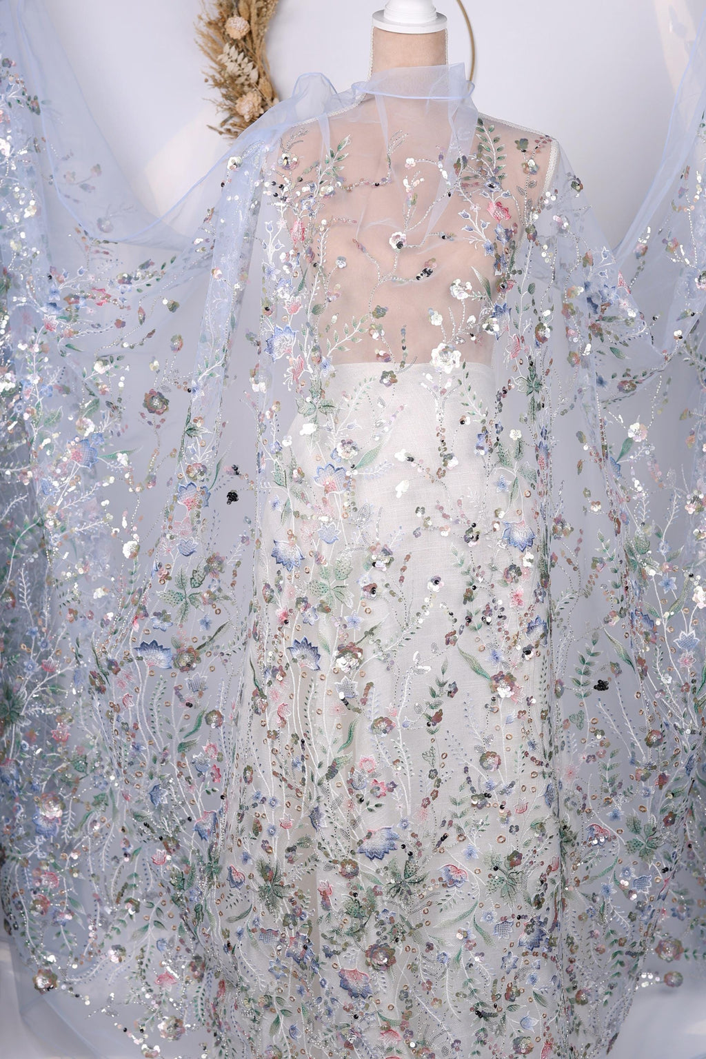 a white dress with a sheer, floral-patterned cape draped over it. The dress appears to be made of sheer fabric, giving it a delicate and elegant appearance. The cape is adorned with a floral design, adding a touch of sophistication to the overall look. The dress and cape are displayed against a white background, which further accentuates their colors and patterns.