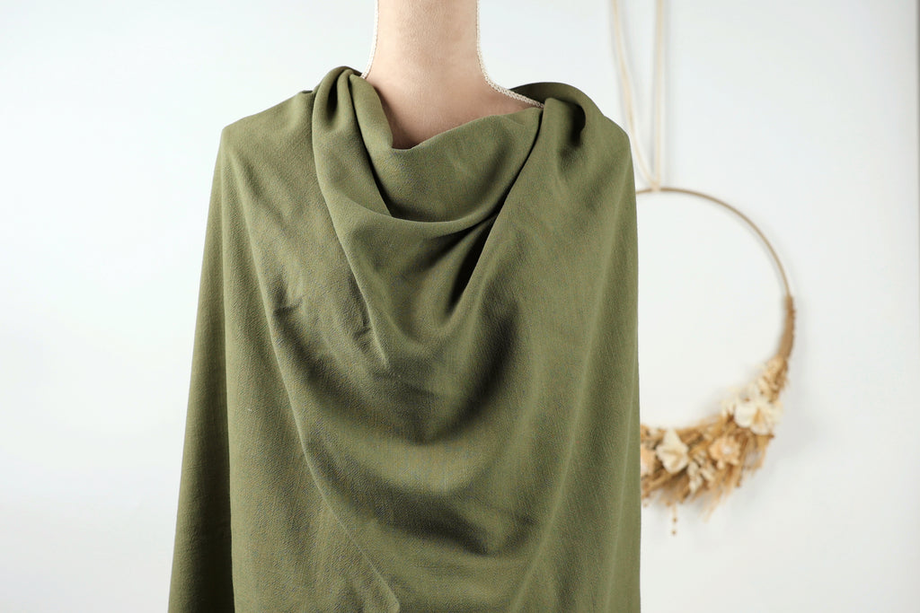 A mannequin wearing a green, rolled-up garment stands in front of a white wall with a wreath hanging on it.