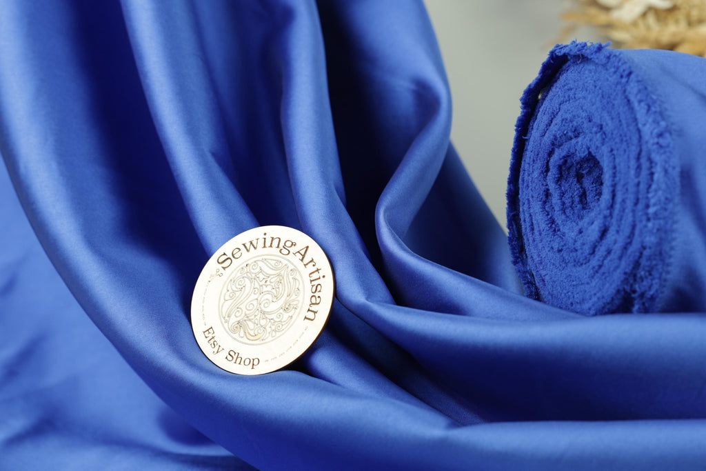 a close-up of a blue fabric with a round metal badge prominently displayed on it, which reads "Sewing Artist" and "Eys Shop".