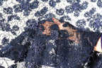 a close-up view of a fabric with a floral pattern, featuring a mix of blue and white colors. The fabric appears to be made of lace or a similar material, with intricate details and a textured appearance.