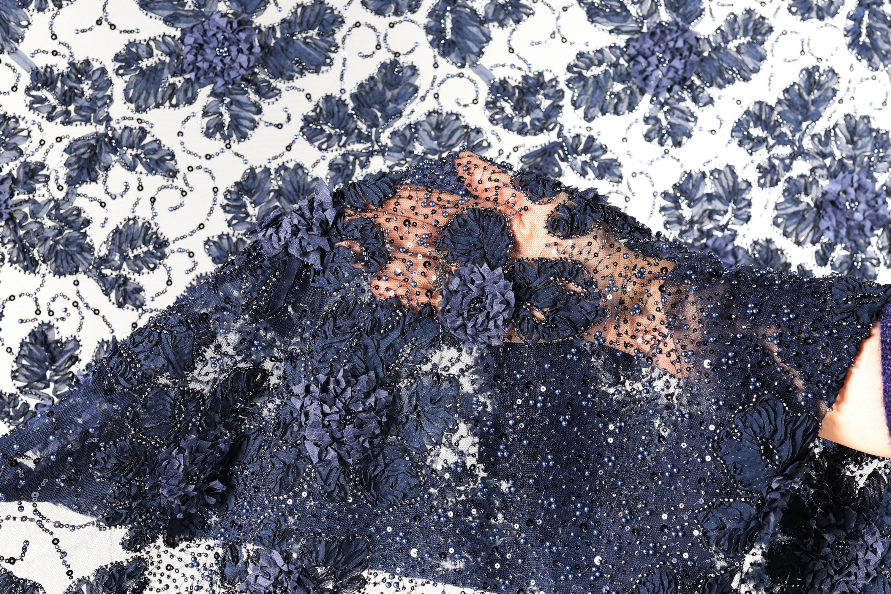 a close-up view of a fabric with a floral pattern, featuring a mix of blue and white colors. The fabric appears to be made of lace or a similar material, with intricate details and a textured appearance.