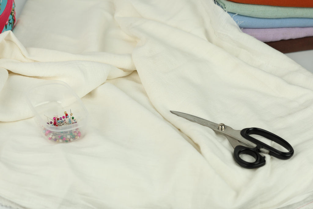 A pair of scissors and a container of pins are placed on a white fabric, suggesting a sewing or crafting project.