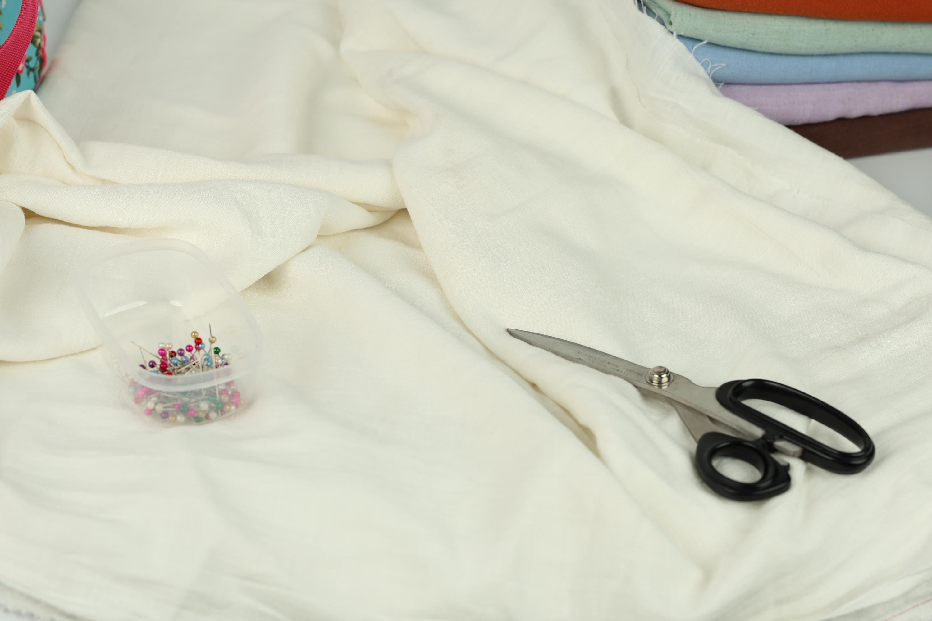 A pair of scissors and a container of pins are placed on a white fabric, suggesting a sewing or crafting project.