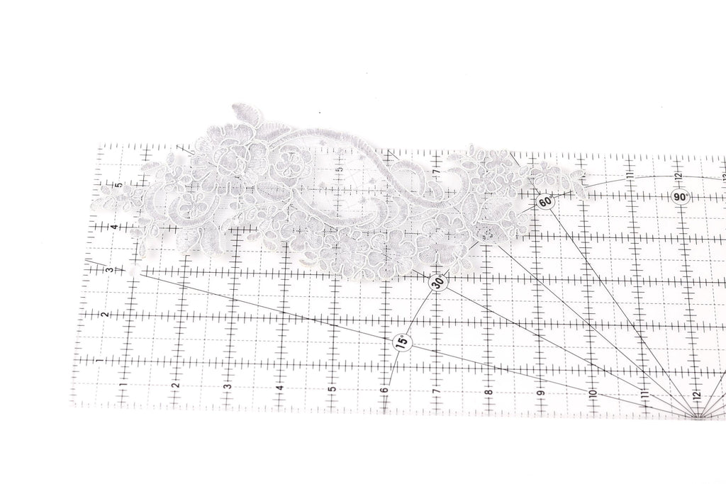 a close-up view of a piece of white lace with intricate floral patterns, placed on a ruler for scale.