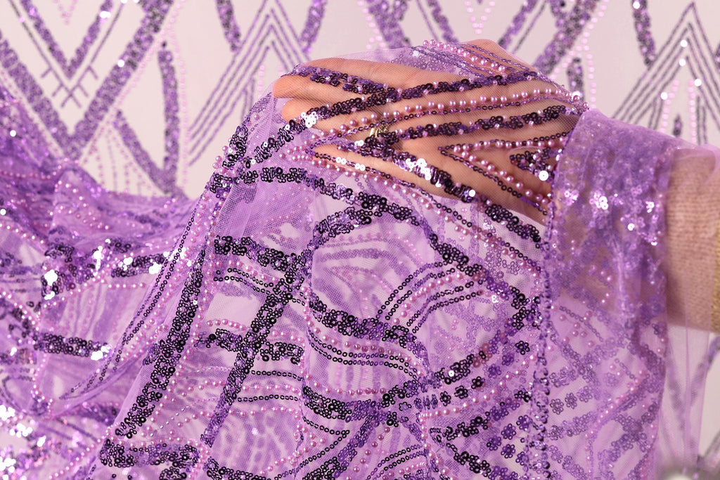 A close-up view of a hand holding a piece of purple fabric adorned with sequins and beads, against a background of a repeating pattern of purple and white geometric shapes.