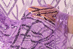 A close-up view of a hand holding a piece of purple fabric adorned with sequins and beads, against a background of a repeating pattern of purple and white geometric shapes.