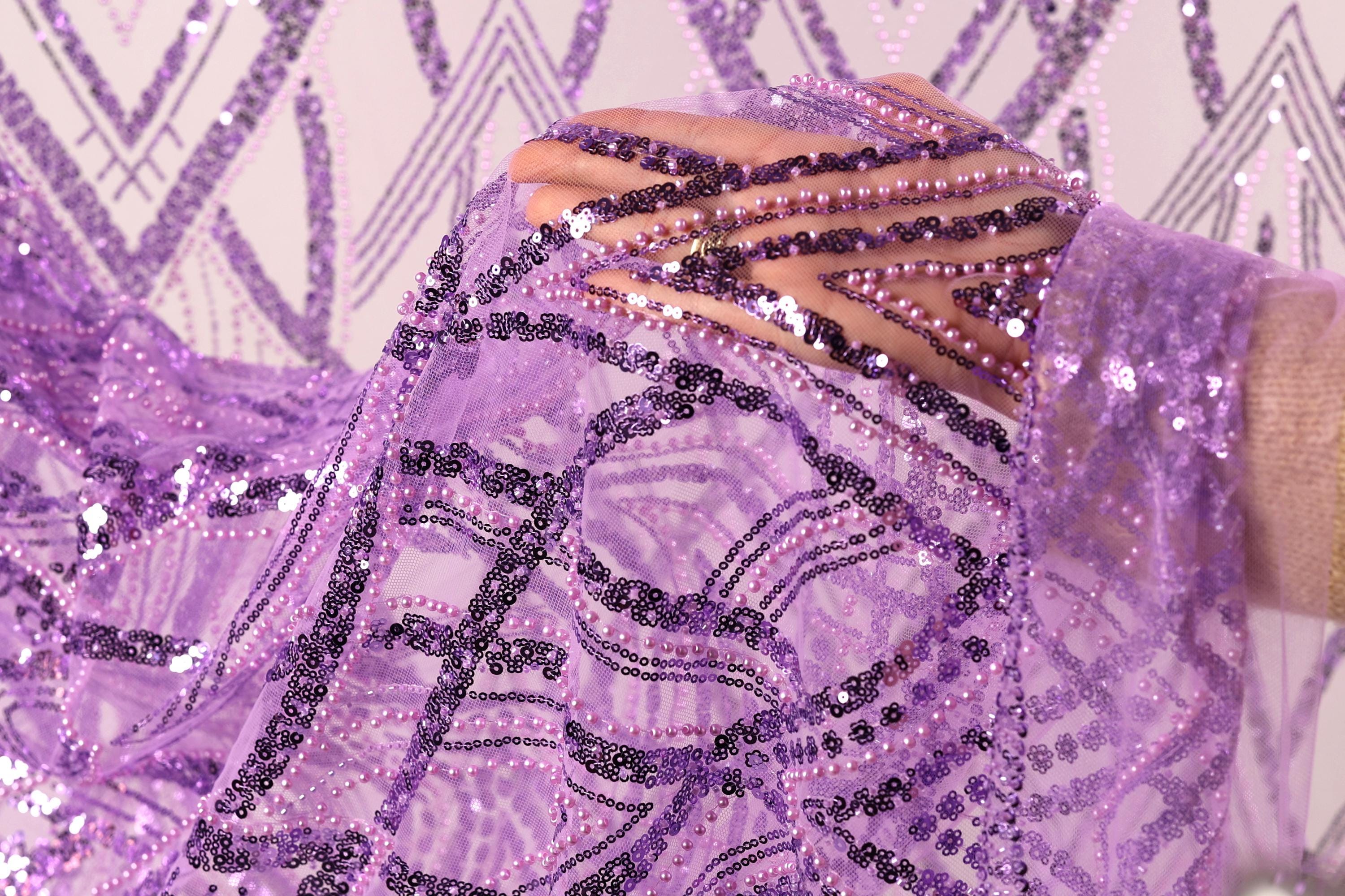 A close-up view of a hand holding a piece of purple fabric adorned with sequins and beads, against a background of a repeating pattern of purple and white geometric shapes.