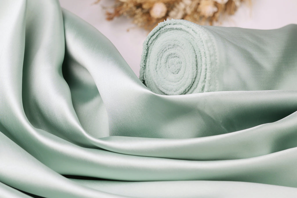A close-up view of a light green satin fabric, possibly silk, with a rolled-up end visible.
