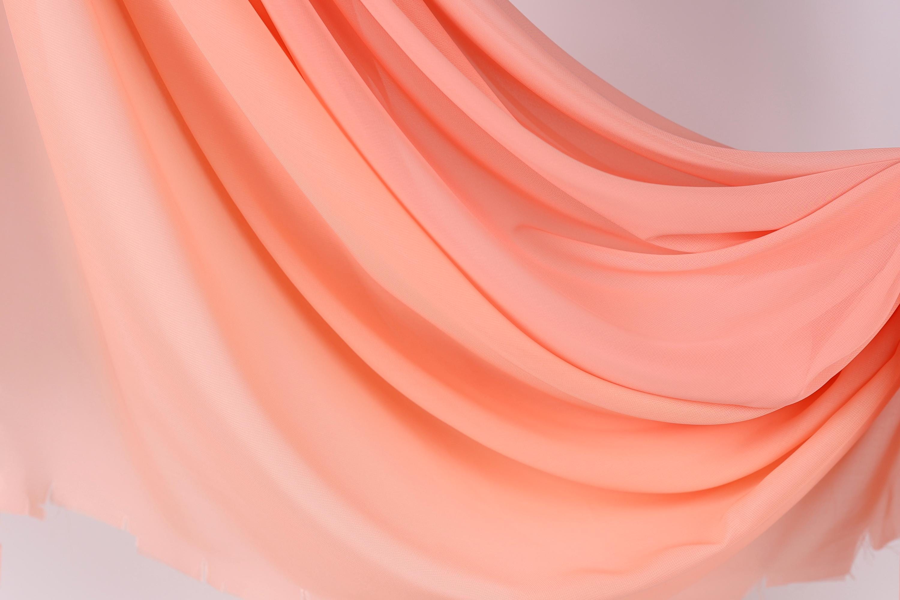 The image is a close-up of a flowing, draped fabric in a soft, peach-colored hue. The fabric appears to be made of a sheer material, giving it a delicate and flowing appearance. The image does not contain any discernible text or countable objects, and the focus is solely on the fabric's texture and color.