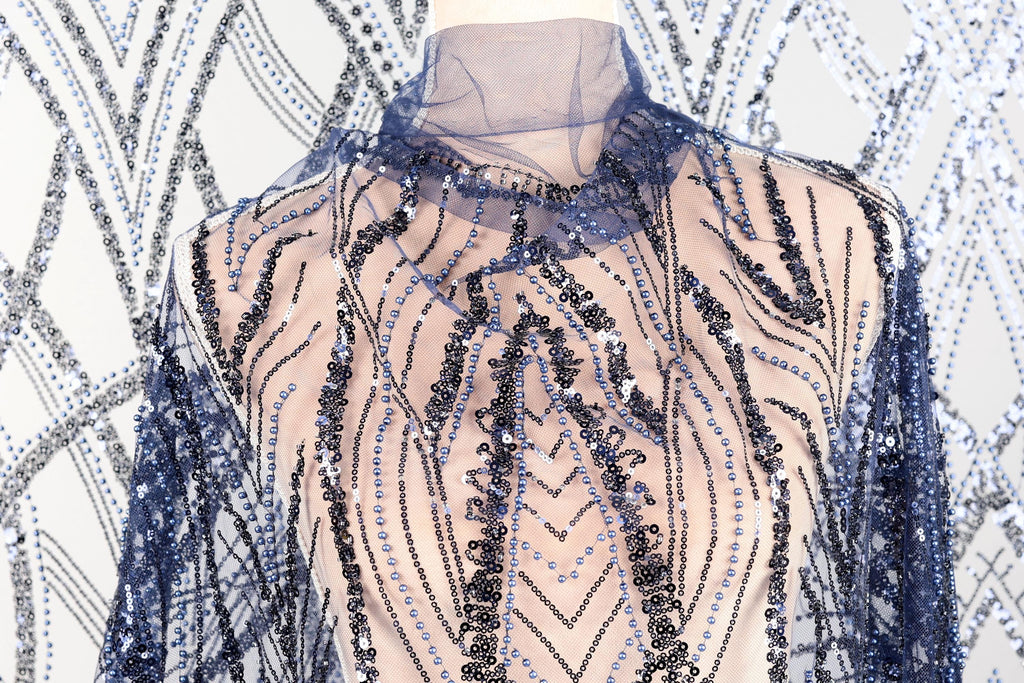 a mannequin wearing a dress with a sheer, sequined bodice and sleeves. The dress has a pattern of blue and black sequins, creating a visually striking and elegant design.