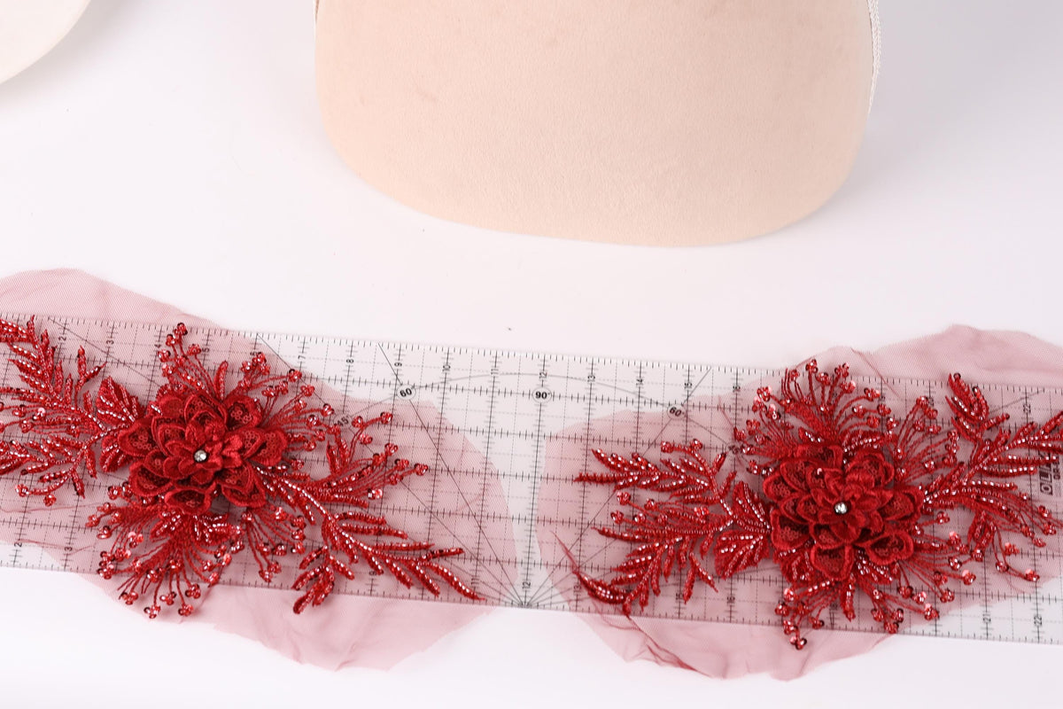 two red floral embellishments with intricate designs, placed on a transparent ruler against a white background.