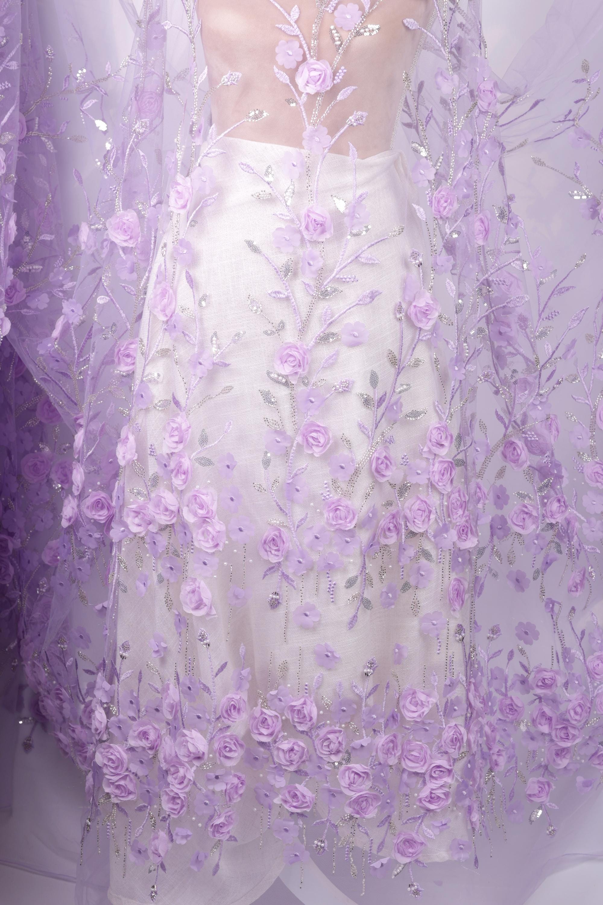 a white dress with a sheer, floral-patterned fabric draped over it. The fabric is adorned with numerous small, delicate purple flowers, creating a visually striking and elegant appearance.