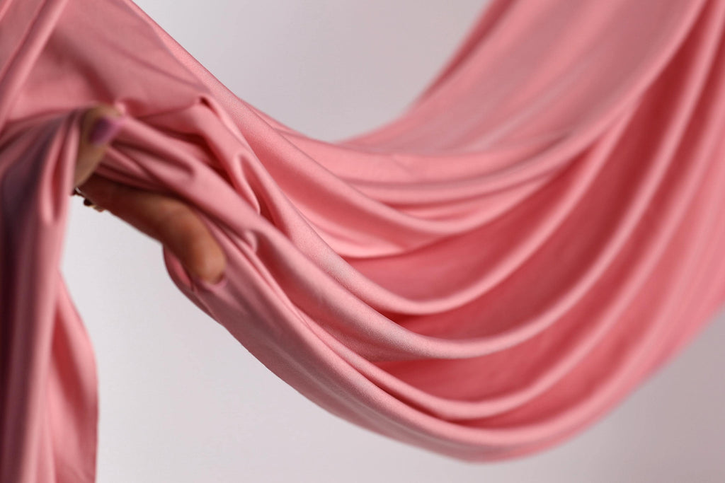 A hand is holding a pink fabric, which appears to be a soft, flowing material. The fabric is being held in a way that it is draped over a surface, creating a sense of movement and elegance.
