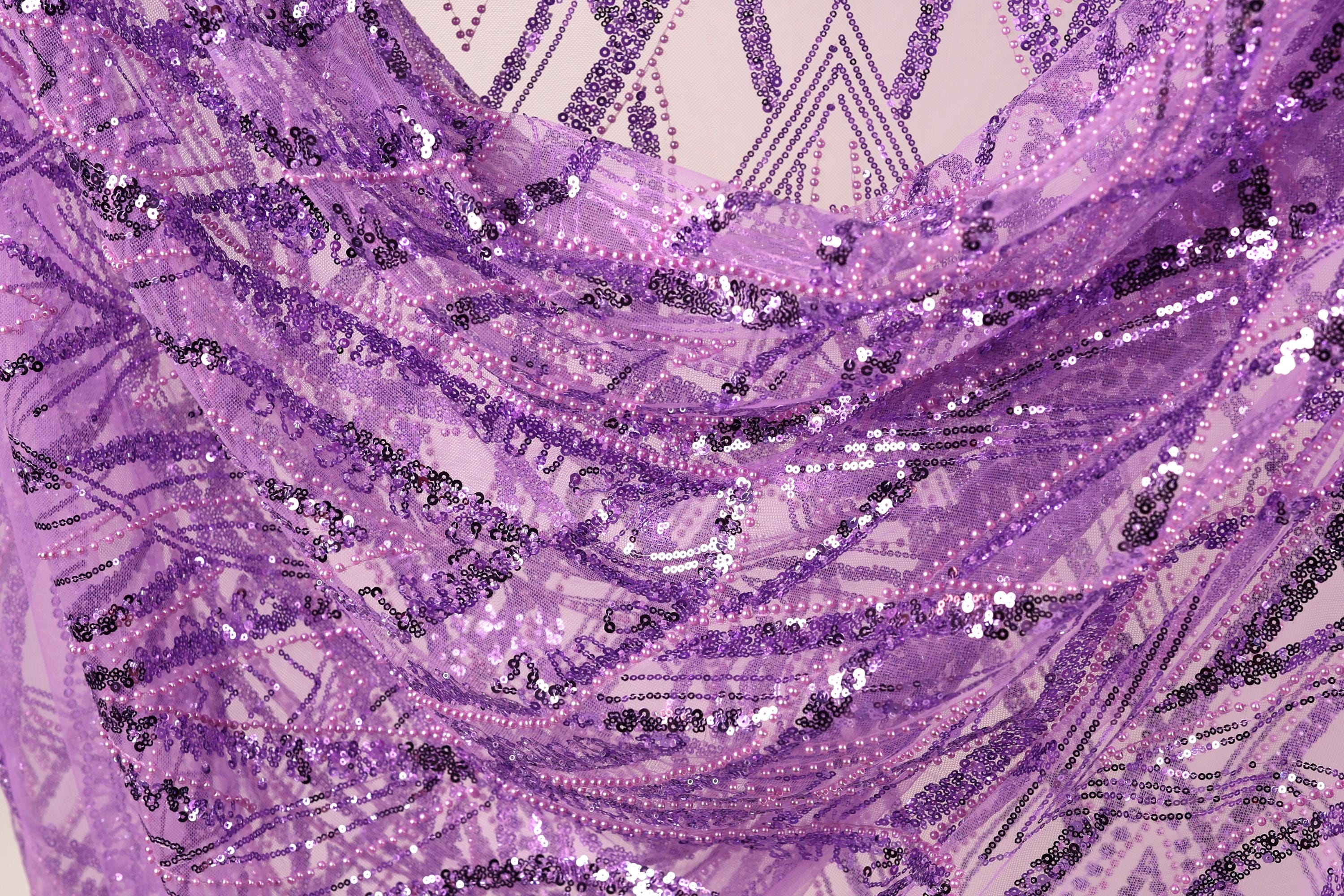 The image is a close-up view of a fabric with a purple and black sequin pattern. The fabric appears to be made of sheer material, with a textured and layered appearance. The image does not contain any discernible text or countable objects, and the focus is on the intricate details of the sequin pattern.