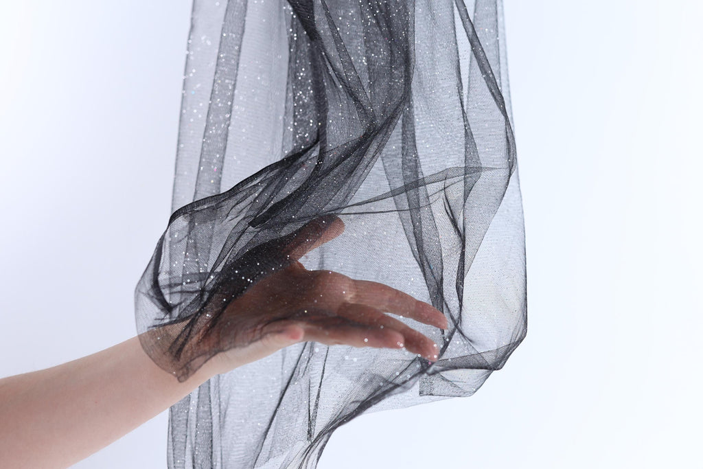 A hand is holding a black, shiny, and textured fabric, which appears to be a sheer or sheer-like material.