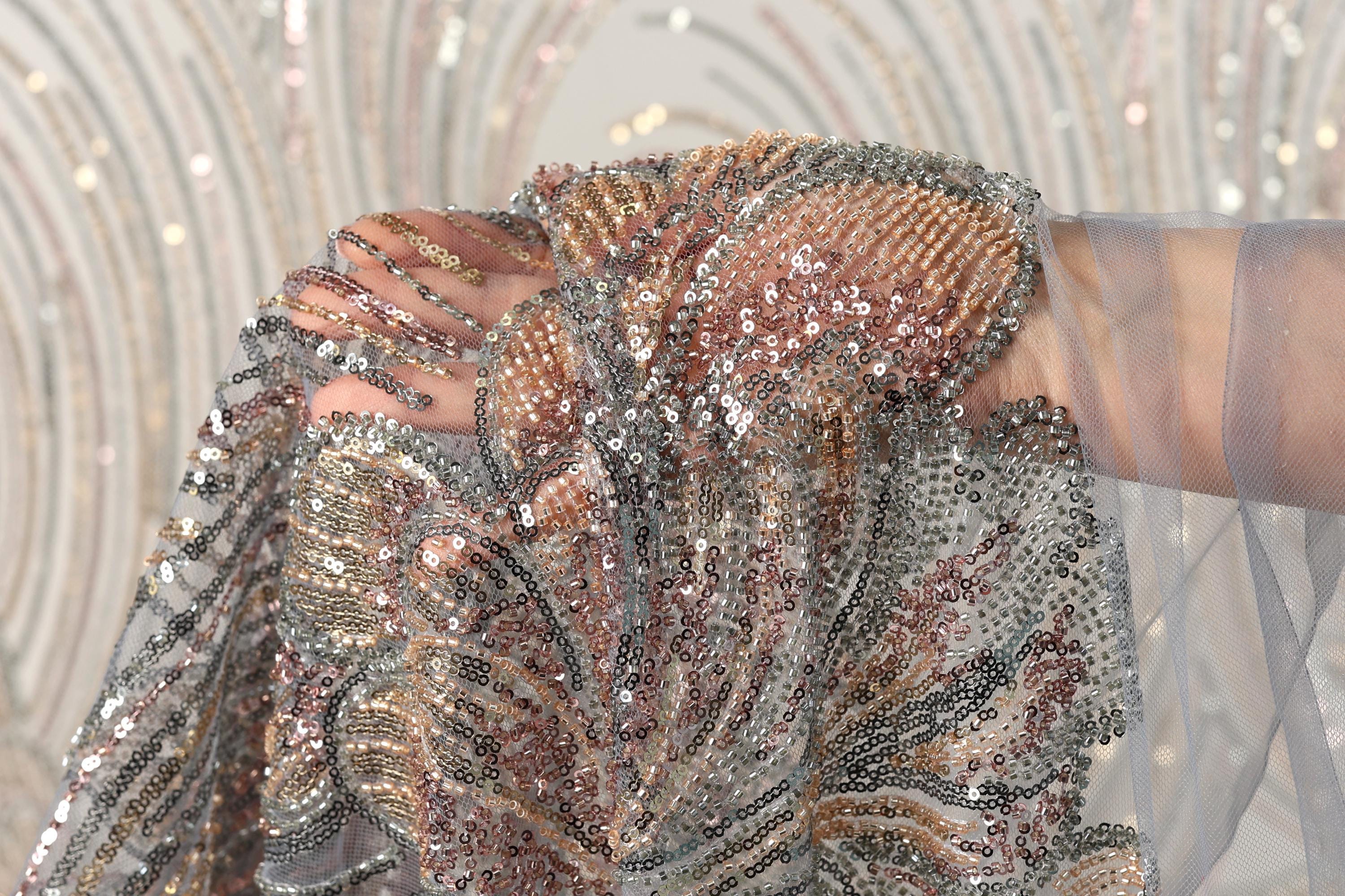 A close-up view of a hand holding a piece of sequined fabric, with the fabric's intricate patterns and shimmering beads visible.
