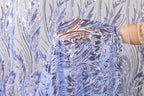 A hand is holding up a piece of fabric with a blue and purple floral pattern, against a background of sequin fabric.