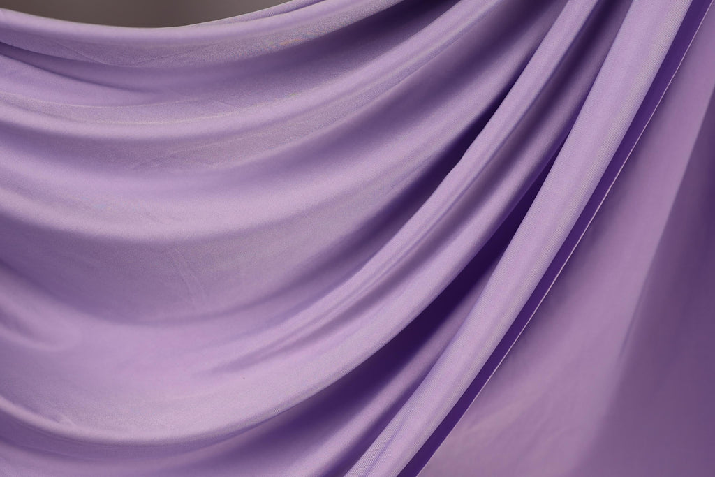 The image is a close-up of a soft, flowing purple fabric, possibly a satin or chiffon material. The fabric has a smooth, shiny texture and is draped in a way that creates a sense of movement and elegance.
