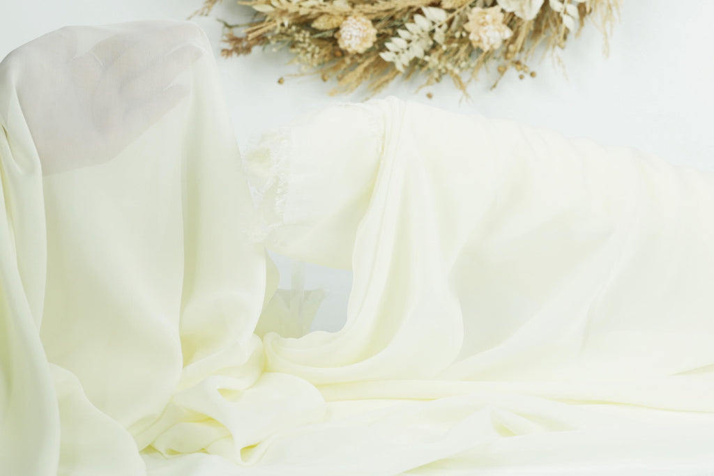 a close-up of a white fabric, possibly a sheet or a garment, with a floral arrangement in the background.